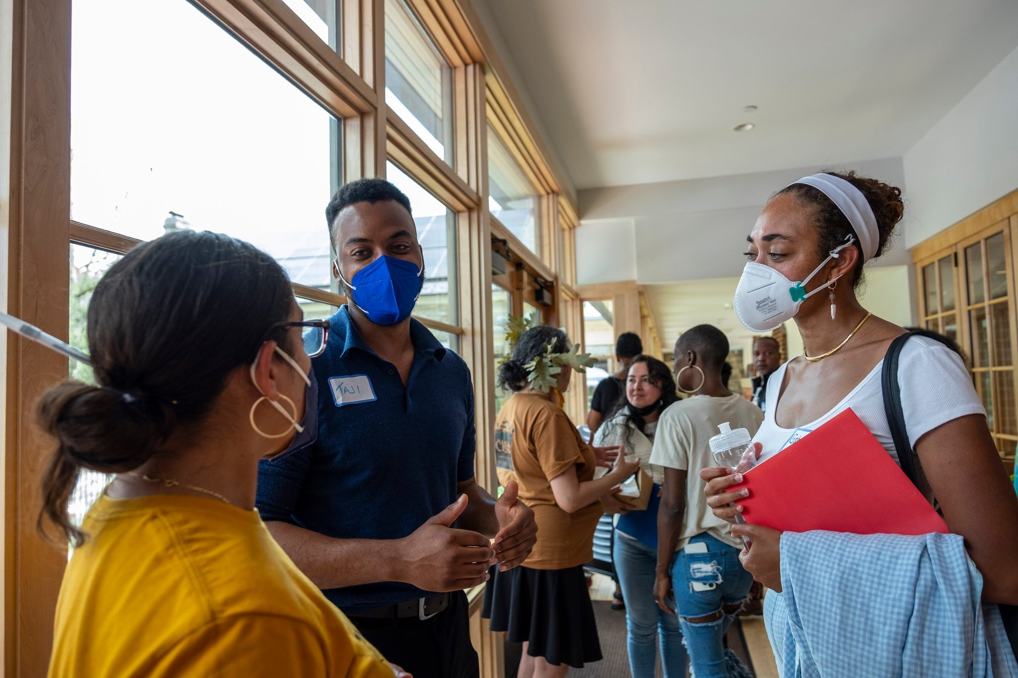 A group of people talk in a hallway. They are all wearing masks and appear to be making friendly conversation. A group of people talk in a hallway. They are all wearing masks and appear to be making friendly conversation.