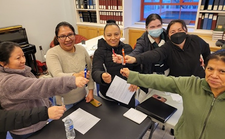 A group of worker owners from Dulce Hogar sit around a table looking happy. They are holding pieces of paper and putting their pens together in the middle.