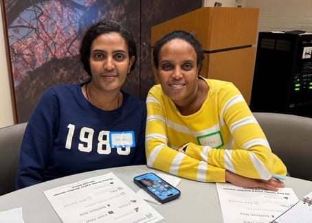 Two people sit at a table and smile at the camera. In front of them on the table, there are worksheets.