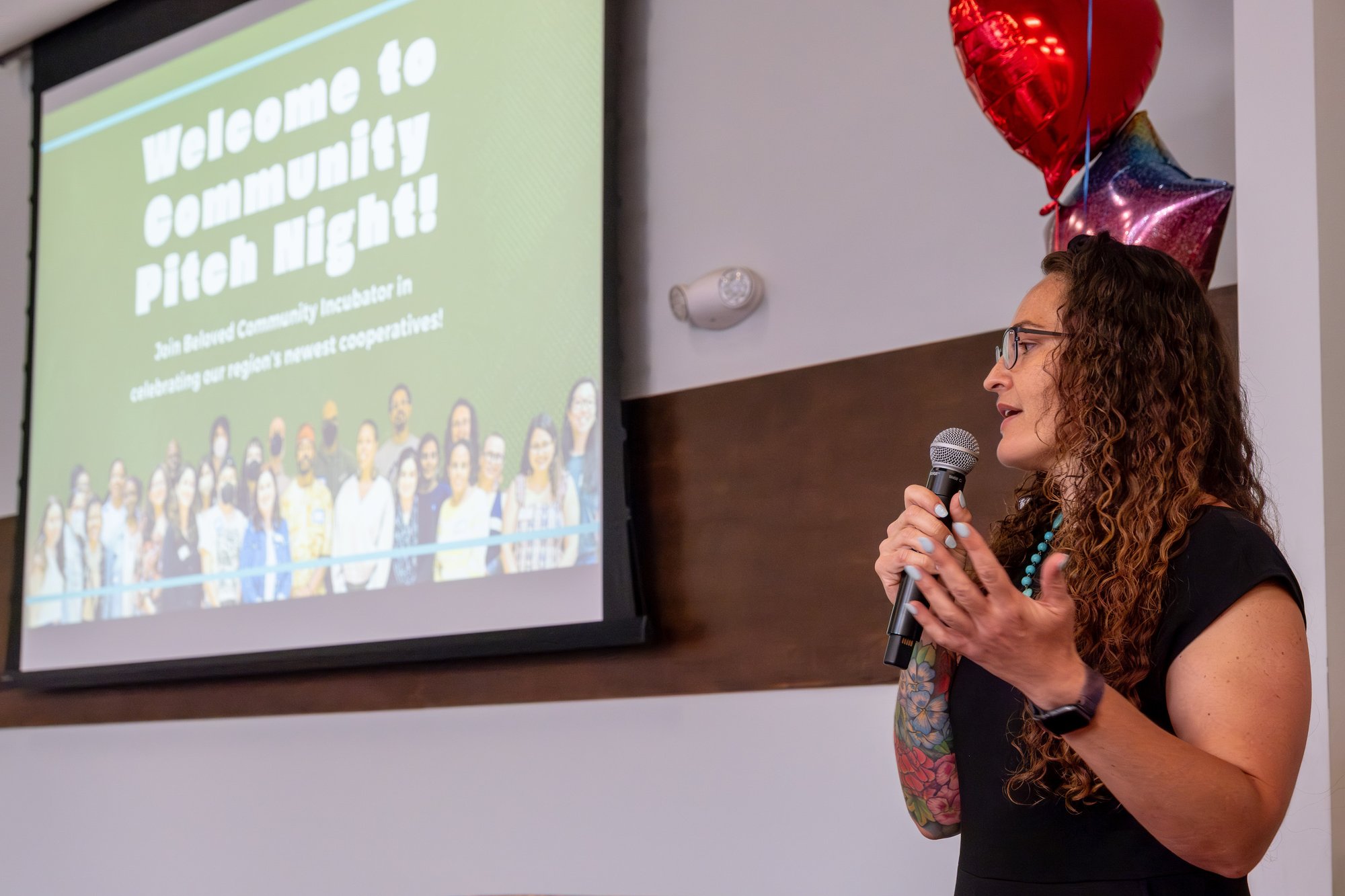 A person stands next to a screen with a slide projected onto it. The person is speaking into a microphone, and the slide reads "Welcome to Community Pitch Night!" with a photo of people on a green background. A person stands next to a screen with a slide projected onto it. The person is speaking into a microphone, and the slide reads "Welcome to Community Pitch Night!" with a photo of people on a green background.