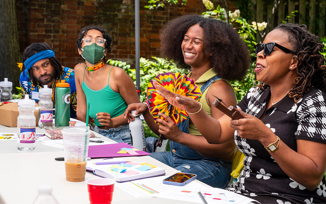 Four people sit next to each other at a table and look happy, they appear to be mid conversation. One holds a brightly colored fan, and all are dressed as though it is hot outside. One person wears a mask. Four people sit next to each other at a table and look happy, they appear to be mid conversation. One holds a brightly colored fan, and all are dressed as though it is hot outside. One person wears a mask.