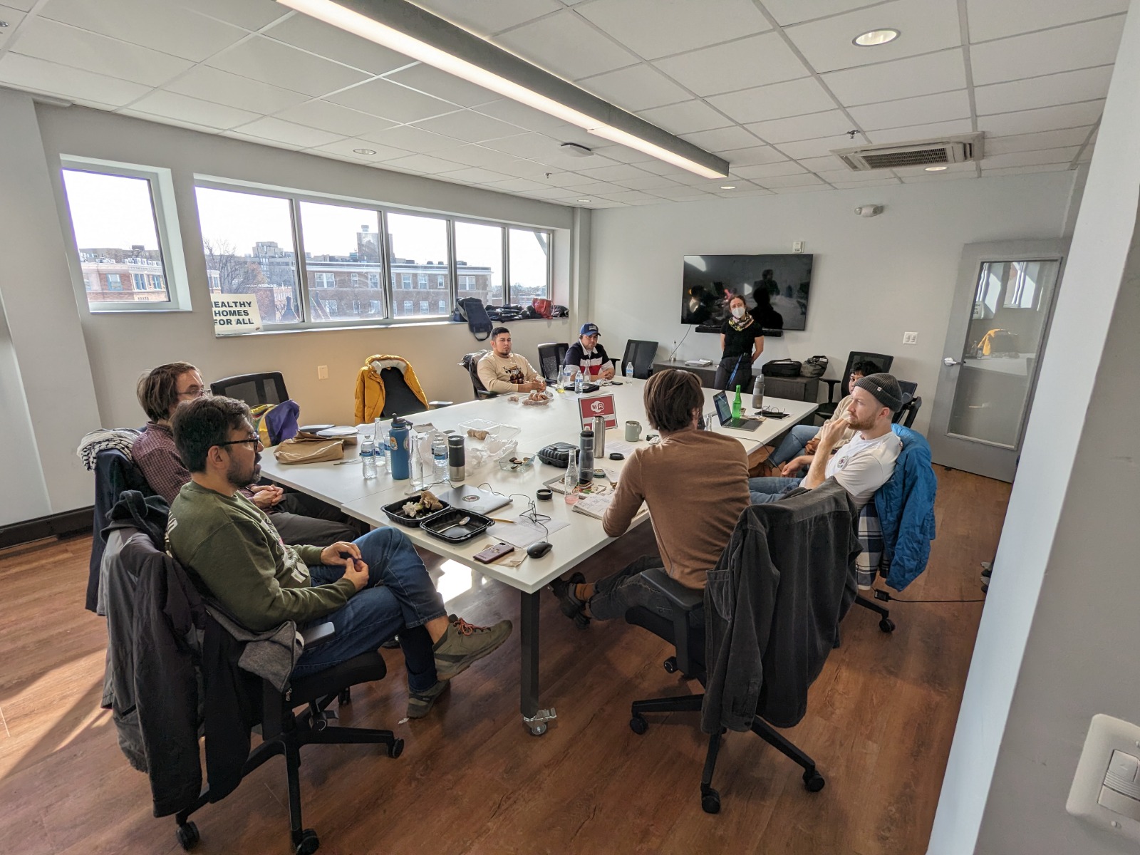 A group of people sit around a table in a conference room. People are dressed casually and appear to have just eaten a meal. Some are masked, others are not. Some laptops are also open in front of people around the table. A group of people sit around a table in a conference room. People are dressed casually and appear to have just eaten a meal. Some are masked, others are not. Some laptops are also open in front of people around the table.
