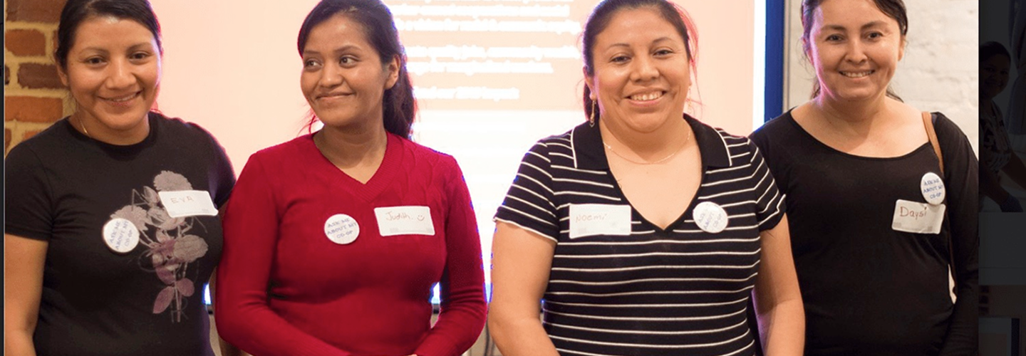 Worker owners of Dulce Hogar smiling at the camera during a presentation in front of a room. Worker owners of Dulce Hogar smiling at the camera during a presentation in front of a room.