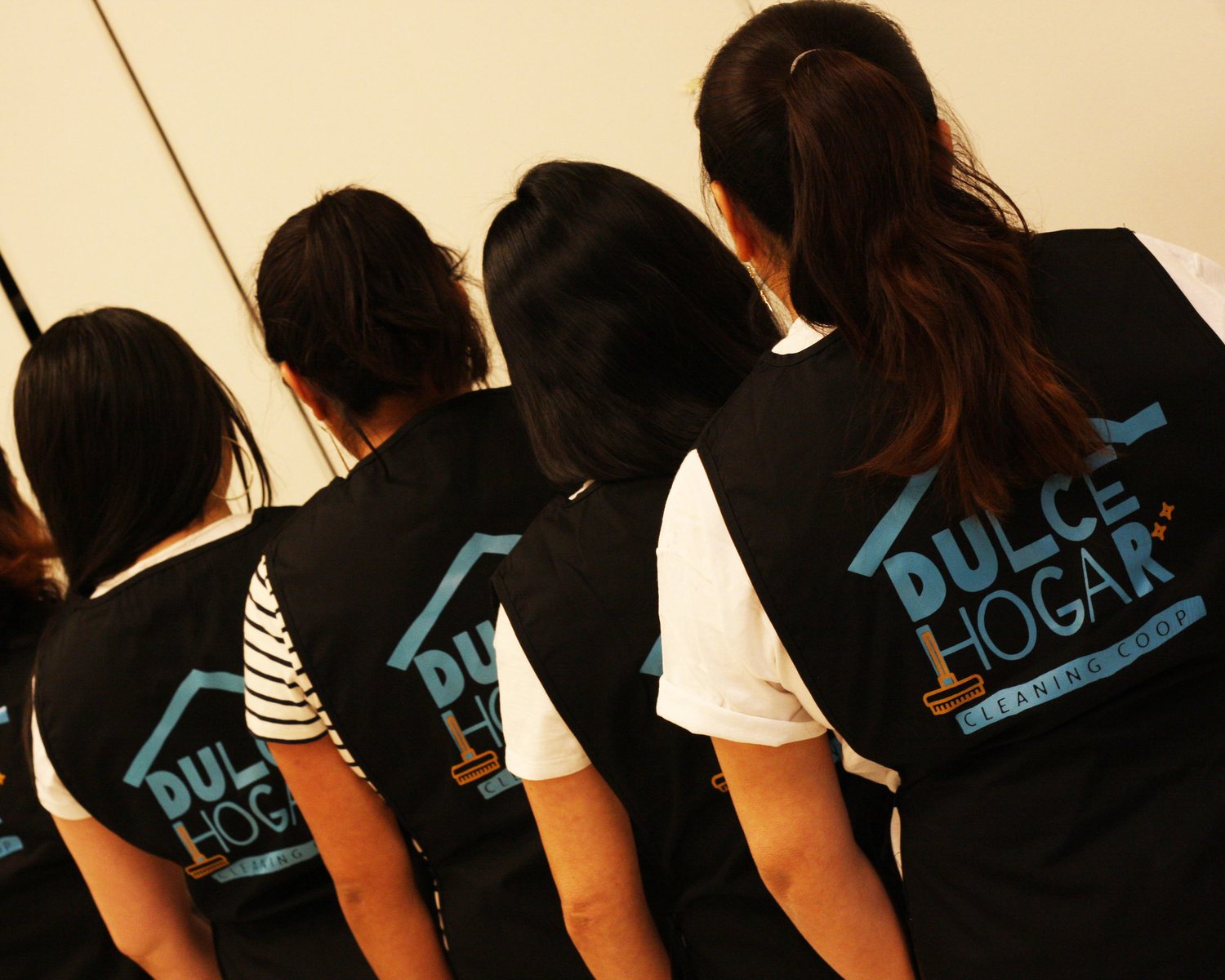 Four people stand with their backs facing the camera near a wall. They wear identical vests, which say "Dulce Hogar Cleaning Co-op" on the back. Four people stand with their backs facing the camera near a wall. They wear identical vests, which say "Dulce Hogar Cleaning Co-op" on the back.