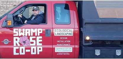A worker owner sits in a new red truck with a large logo for Swamp Rose Co-op on the side. Another sign on the truck reads "Ecological Landscaping."